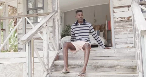African american man sitting on front steps of wooden beach house relaxing in Stock Footage 337699702