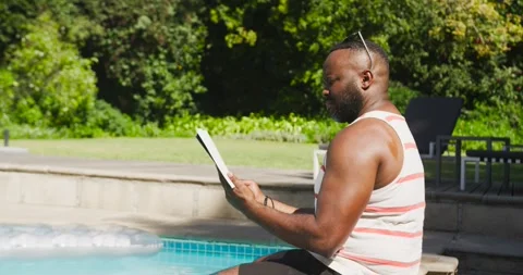 African American man sitting poolside reading book, flipping pages and adjusting Stock Footage 340882062