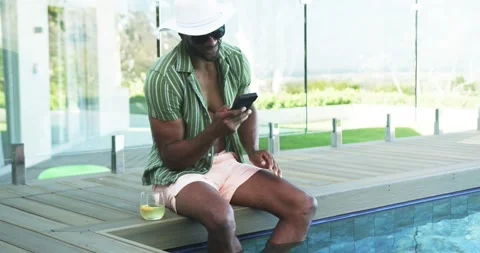 African American man sitting poolside on deck in striped shirt, tapping Stock Footage 340969429