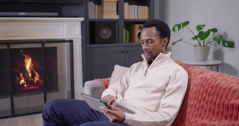 African American man sitting on sofa seeing tablet, tapping, pointing at fire, Stock Footage 339231489