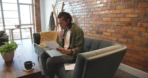 African American man sitting on sofa in loft, reviewing documents, typing on Stock Footage 339959393