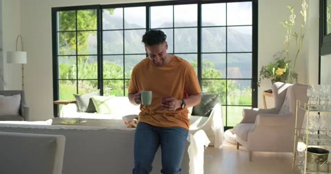 African American man sitting on sofa wearing orange tee, reading phone text, Stock Footage 344851086