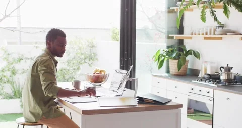 African american man sitting at table in kitchen, using laptop and taking notes Stock Footage 338433863