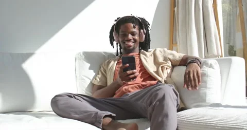 African American man sitting on white sofa with headphones holding phone and Stock Footage 341321069