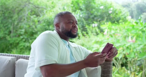 African American man sitting on wicker sofa on porch holding pink phone reading Stock Footage 337933155
