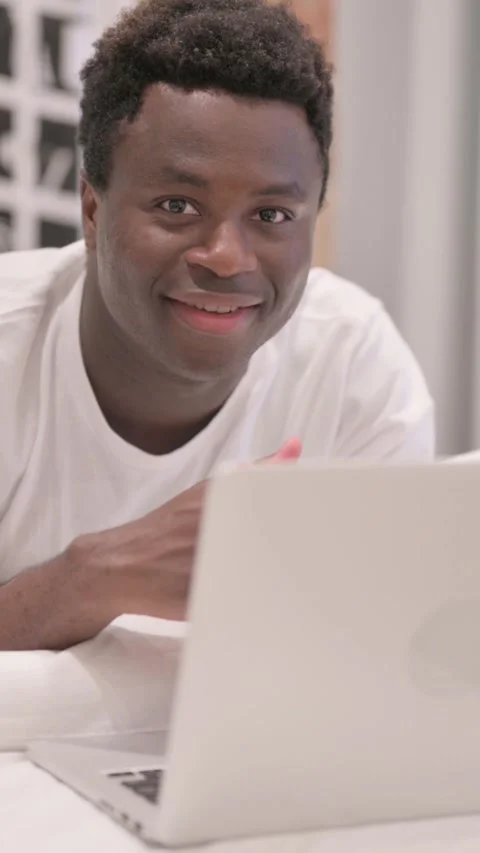 African American Man Smiling at Camera while Working on Laptop while Lying on 스톡 동영상 328568254