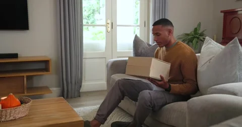 African American man on sofa checking phone, placing box on table beside woven Stock Footage 342363385