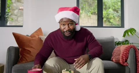 African American man on sofa lifting gift box, revealing surprise, sharing joy Stock Footage 319779941