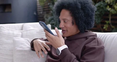 African American man taking call, chatting, scrolling phone on patio sofa, Stock-Footage 345607909
