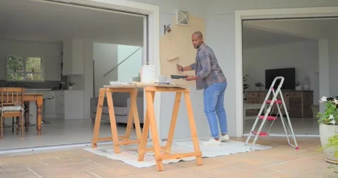 African American man touching-up wall patch on porch using roller; phone Stock Footage 339321130