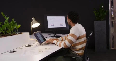 African American man typing, studying monitor charts and writing notes at office Stock Footage 345589257