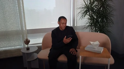 African American Man using app on cell phone while sitting on sofa. 스톡 동영상 108601167
