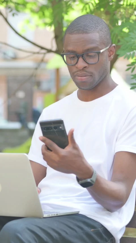 African American Man Using Laptop and Phone while Sitting Outdoor, Vertical Stock Footage 319028014