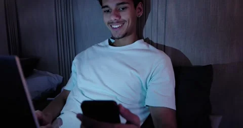 African American man using laptop in bedroom, pausing, picking up smartphone and Stock Footage 342084629