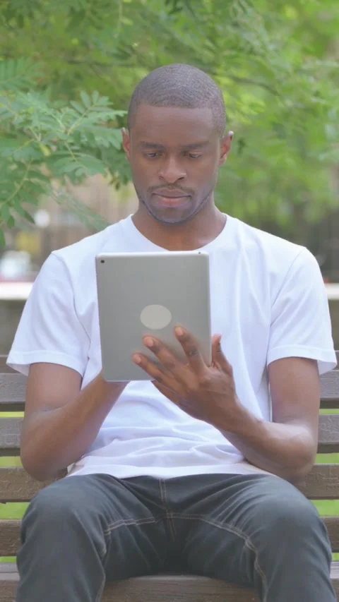 African American Man Using Tablet while Sitting on Bench in Park, Vertical Video 스톡 동영상 319025912
