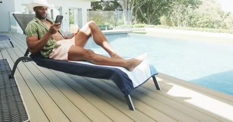 African American mature man lying on lounger by pool, scrolling phone, sipping Stock Footage 337625035