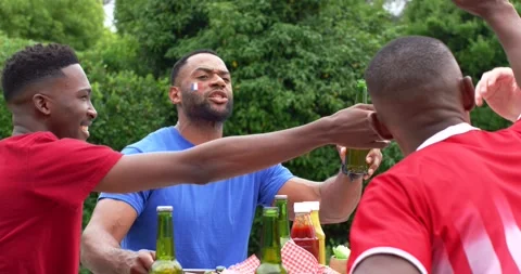 African American men at yard table red shirt reaching bottle blue shirt offering Stock Footage 335987427