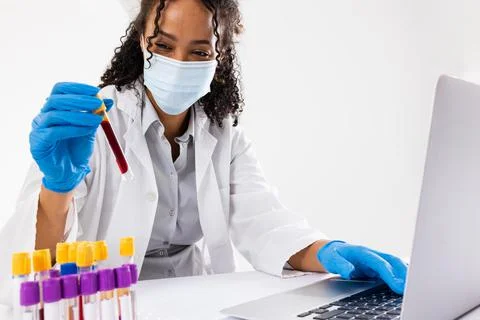 African american mid adult woman wearing mask holding test tube and using laptop Stock Photos