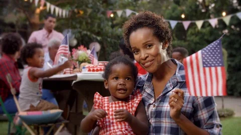 African American mother and baby girl wave flag at 4th July party Stock Footage