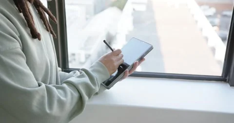 African American nonbinary in hoodie using stylus on tablet drawing at window, Stock Footage 344693501