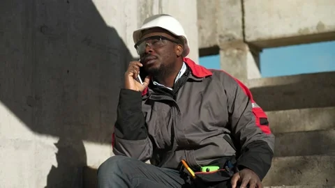 African american worker talks on smartphone. Builder sits on concrete stairs Stock Footage 201199280