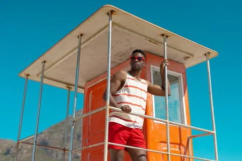 African american young man wearing sunglasses standing on lifeguard hut against Stock Photos