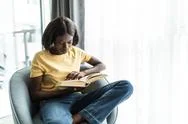 African American Young Woman Reading Book And Sitting On Chair At Home. Peopl Stock Photos