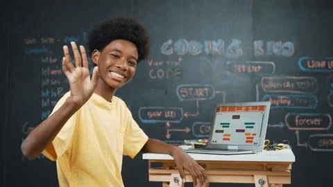 African boy use laptop to code program and turn around to wave hand. Edification Photos