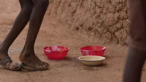 African boy washes spoon in bowl of water squatting near a clay building. Slow Video stock 311334792