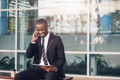 African businessman laughing while using his phone and digital tablet Stock Photos