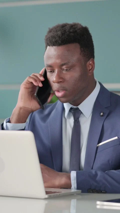 African Businessman Talking on Smartphone while using Laptop in Office, Vertical Vídeos de archivo 339574695