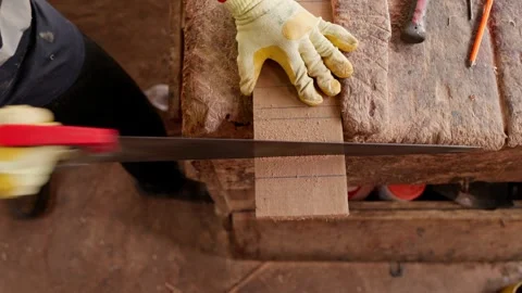 African carpenter in work gloves saws wooden plank with hand saw on workbench Stock Footage 346733717