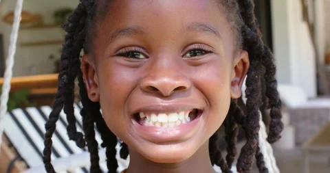 African child smiling while camera gaze widening grin on porch swing, braids Stock Footage 336685505