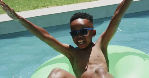 African child in trunks with shades sitting in green ring at backyard pool Stock Footage 344105398