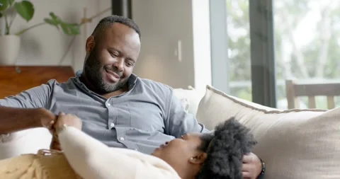 African couple sitting on light-colored couch, man looking down, reaching, Stock Footage 338337374