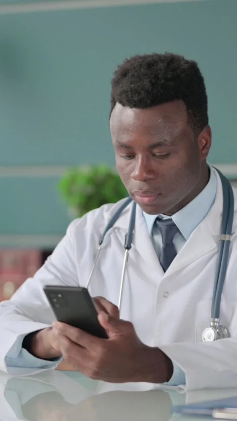 African Doctor using Smartphone while Working on Laptop in Office, Vertical Stock Footage 339575283