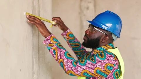 African engineer in hard hat, vest checks wall length with tape on site Video stock 346257515