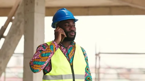 African engineer in hard hat, vest talks on phone at site Stock Footage 346257563