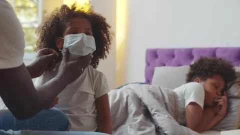 African father putting safety mask on little daughter with sick son lying in bed Stock Photos