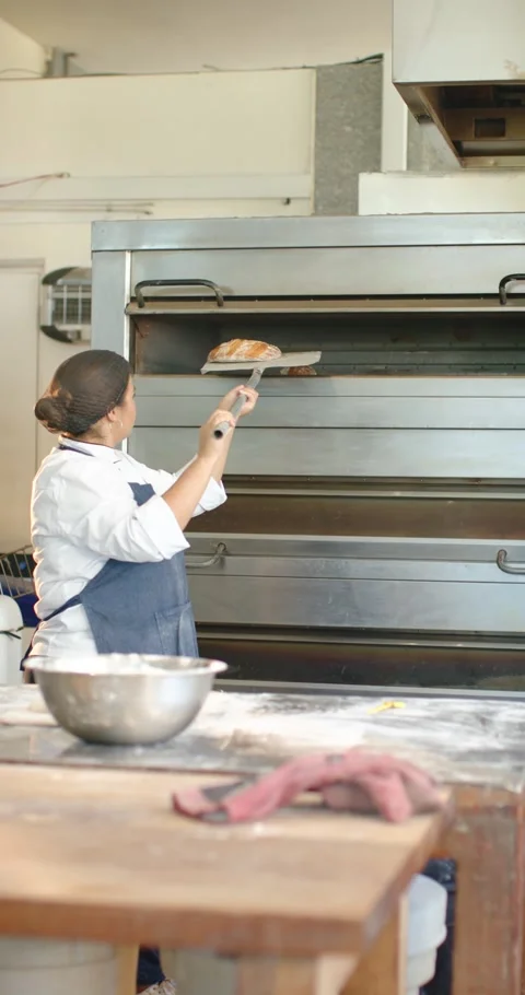 African female baker in apron checking loaf, opening oven, sliding peel to Video stock 345397129