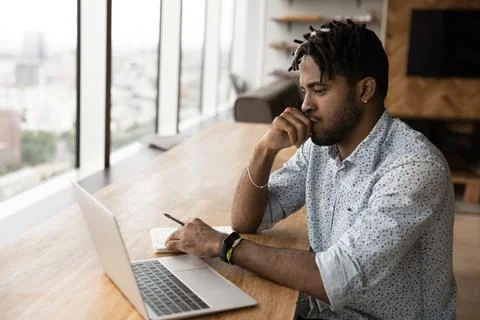 African guy sit at table looks at laptop consider task Foto stock