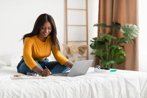 African Lady Learning Online Using Laptop Taking Notes In Bedroom Stock Photos