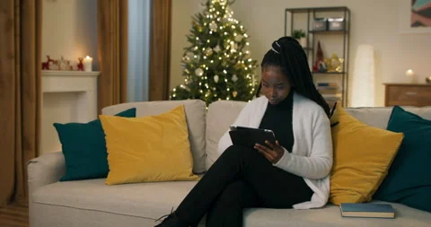 An African-looking student sits on sofa, holding tablet and ordering Christmas Stockbeeldmateriaal 200854466