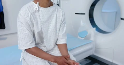 African man clasping rubbing hands while sitting on radiology table wearing gown Stock Footage 344333609