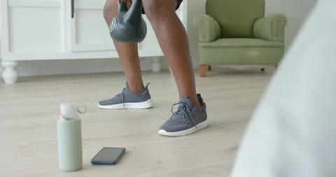 African man doing kettlebell squats on living room floor in shorts after phone Stock Footage 345037016