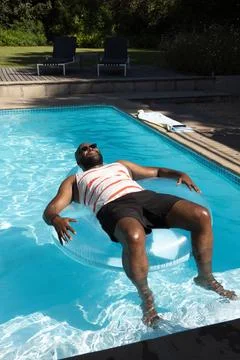 African man floating on clear inflatable ring in backyard pool, wearing striped Stock Photos