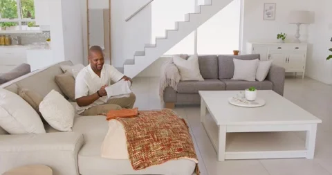 African man inspecting and folding garments while sitting left on sofa, stacking Stock Footage 339416082
