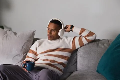 African man leaning back on sofa at home, wearing cream sweater, headphones, Stock Photos