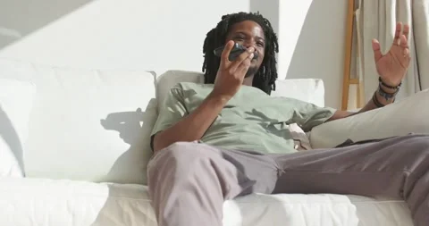 African man leaning on white sofa holding smartphone, speaking and smiling in Stock Footage 342023543