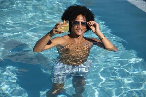 African man in patterned trunks waist-deep in pool, adjusting sunglasses and Stock Photos